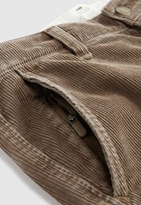 Tessuto a coste marrone con una texture a righe, dotato di una tasca con zip e chiusura a bottone. Il design include dettagli di cucitura sui bordi.