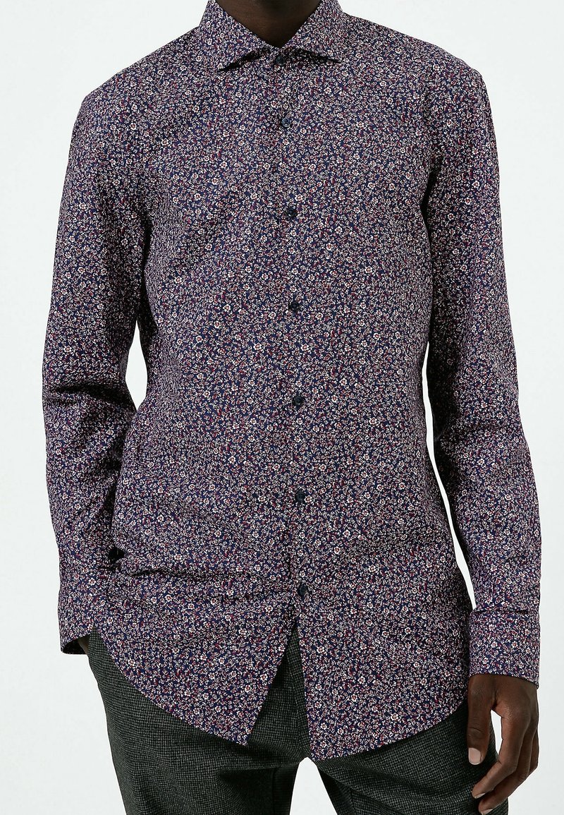 Manskjorta i mörkblått med blommönster, med knapplist, långa ärmar och en rundad fåll. Slät tygtextur.
