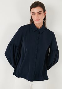 Camicia azzurra scuro con colletto, caratterizzata da maniche a sbuffo e una texture morbida e liscia. Bottoni frontali e orlo arrotondato.