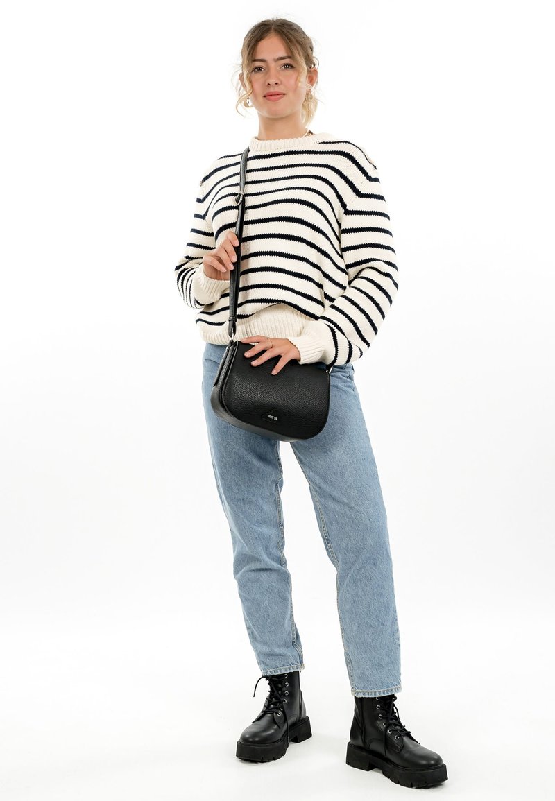 Frau trägt weißen und schwarzen gestreiften Pullover, hellblaue Jeans, schwarze klobige Stiefel und trägt eine schwarze Schultertasche, posiert vor weißem Hintergrund.