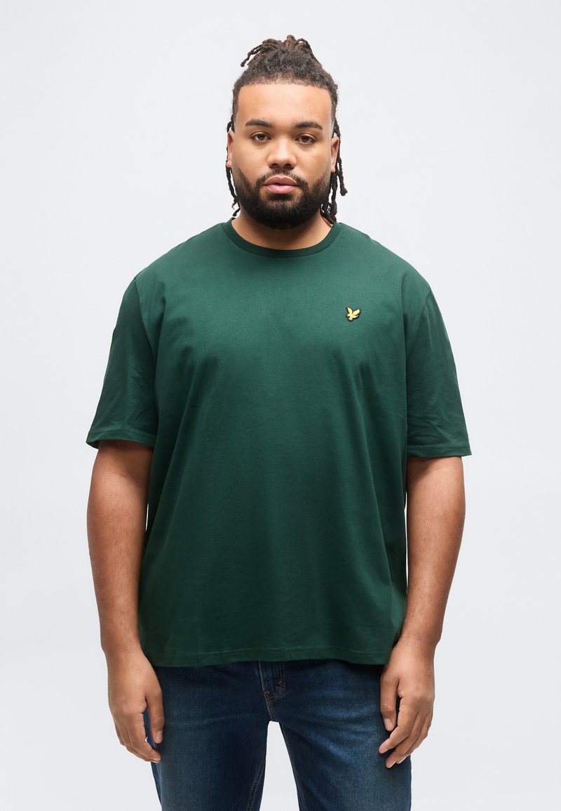 Άνδρας με πλεγμένα μαλλιά και γένια, που φοράει πράσινο σκουρόχρωμο μπλουζάκι με μικρό κίτρινο πουλί ως λογότυπο και μπλε τζιν, που στέκεται μπροστά σε μονόχρωμο φόντο.