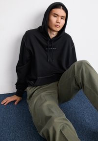 Crna hoodie s kapuljačom i vezicom, u kombinaciji s maslinastozelenim širokim hlačama. Tekstuiran materijal s vidljivim šavovima.