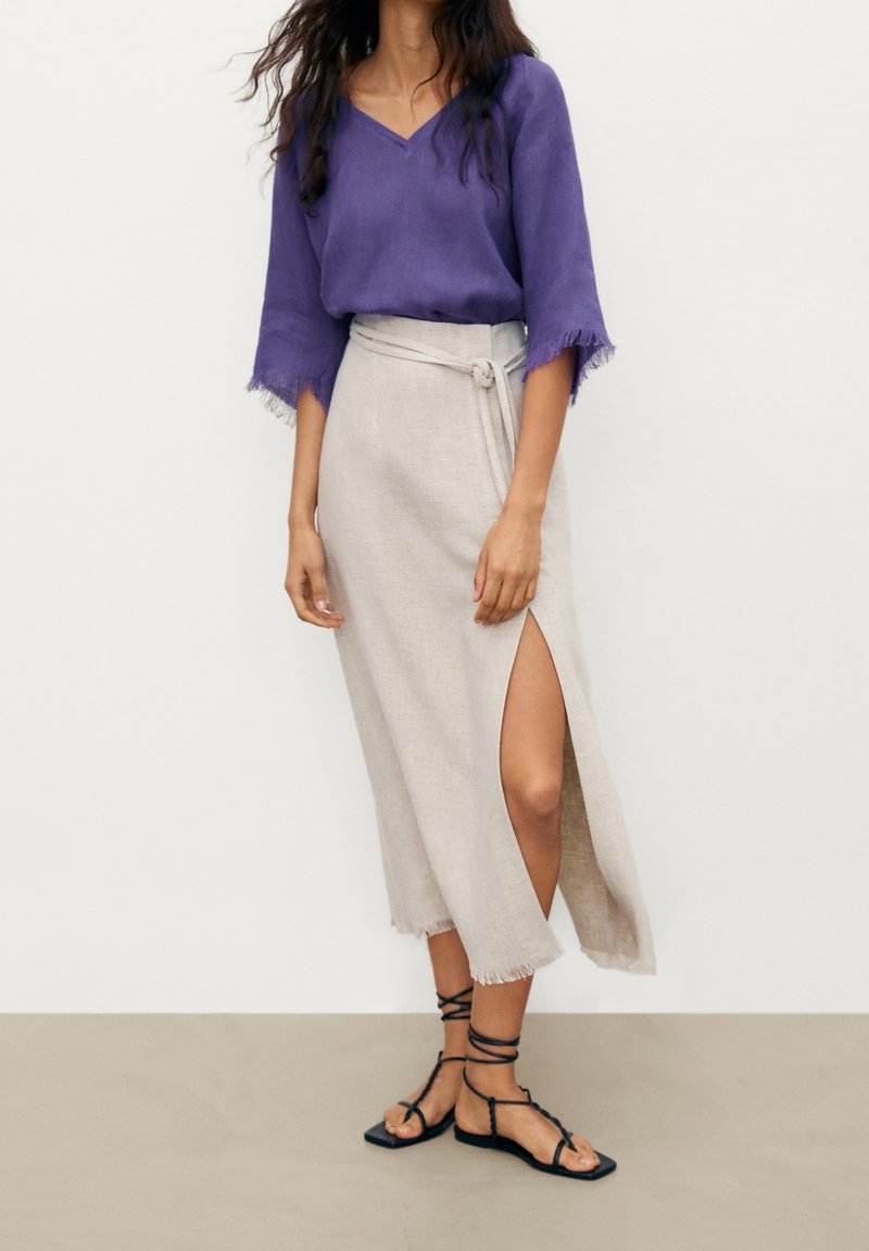 Blouse en lin violet avec manches effilochées, jupe portefeuille en lin beige avec fente latérale, et sandales à lanières noires sur un fond neutre.
