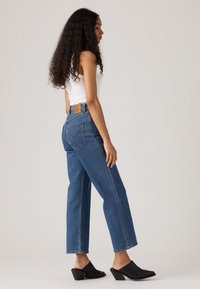 Donna con lunghi capelli ricci, vestita con una canotta bianca senza maniche, jeans blu a vita alta e mule nere con tacco, in piedi di lato contro uno sfondo neutro.