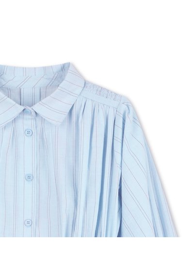 Ανοιχτό μπλε πουκάμισο με γιακά, κάθετες ρίγες με κόκκινες λεπτομέρειες, κλείσιμο με κουμπιά και μανίκια με πιέτες.