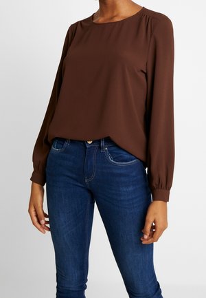 Personne portant un chemisier marron à manches longues rentré dans un jean bleu taille haute avec des coutures visibles et une fermeture à boutons.