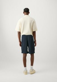 Polo giallo chiaro a maniche corte con tessuto tessurizzato abbinato a pantaloni corti blu navy. Modello che indossa sneakers beige e calze bianche.