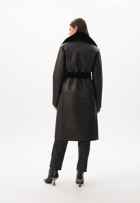 Černý kožený kabát s širokým límcem, páskem v oblasti pasu a dlouhými rukávy; hladká textura a minimalistický design; nošený přes potištěné tmavé kalhoty.