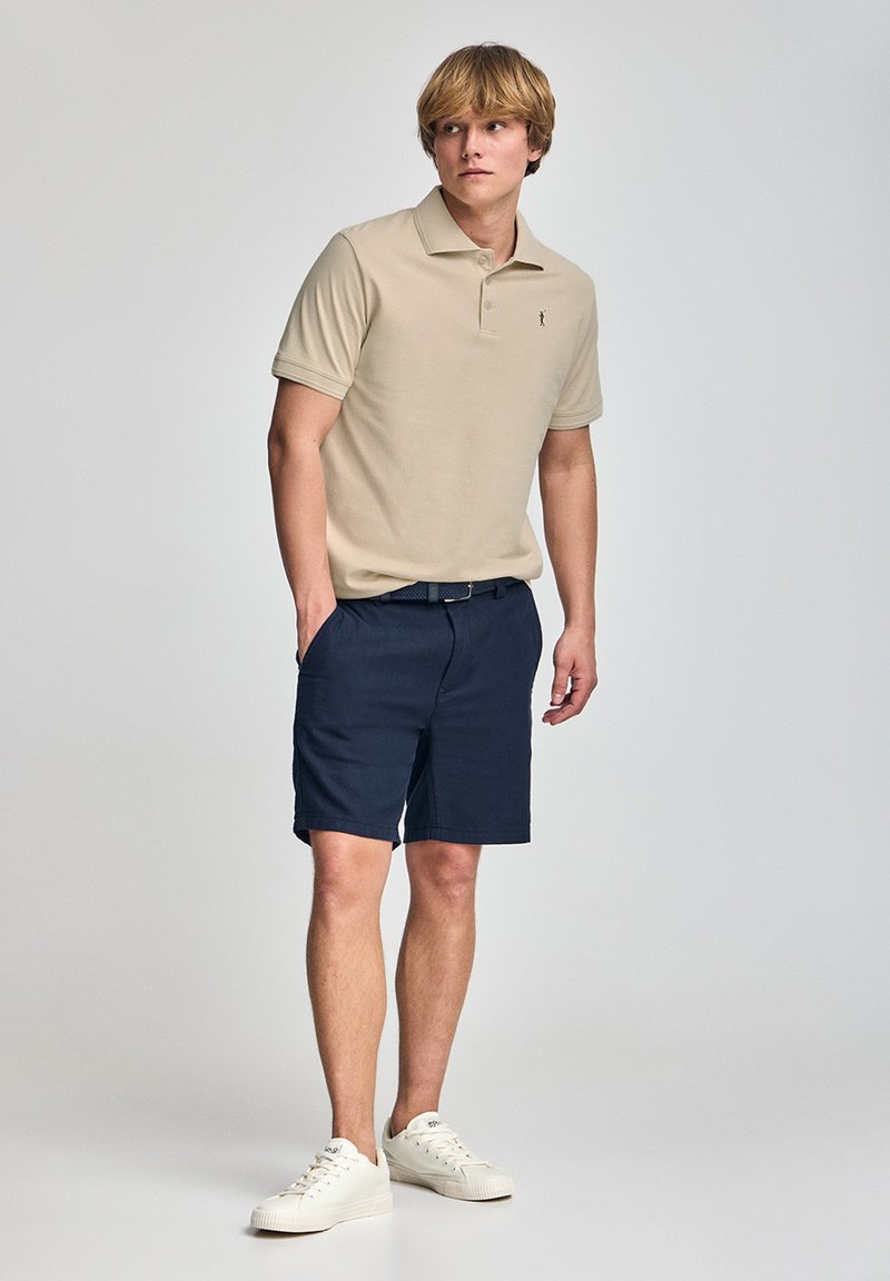 Camisa polo beige con cuello y logo, combinada con pantalones cortos azul marino. El modelo lleva zapatillas blancas, con las manos en los bolsillos y una postura recta.