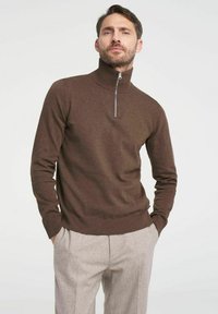 Brauner Pulli mit Reißverschluss und Rippenbündchen am Saum, ausgestattet mit einem hohen Kragen und einem silbernen Reißverschluss. Getragen mit hellgrauen Hosen.