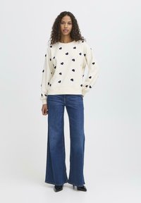 Maglioncino crema con motivi a cuore blu, polsini e orlo a coste; abbinato a jeans larghi blu e scarpe nere. Tessuto liscio.
