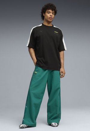 Jonge man staand met handen in de zakken, gekleed in een zwart-wit shirt met korte mouwen, wijde groene broek en zwart-witte schoenen tegen een grijze achtergrond.