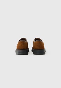 Un par de zapatos casuales de ante marrón con suelas gruesas negras, vistos desde la parte posterior sobre un fondo claro.