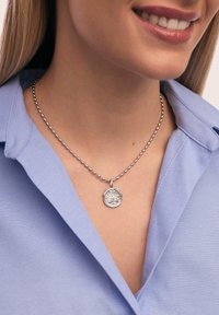 Collier en perles argentées avec un pendentif rond présentant un motif d'arbre, porté sur une chemise col bleu clair.