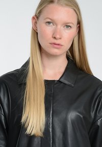 Sort læder skjortejakke med krave, synlige sting og en glat overflade. Modellen har langt, glat blond hår.