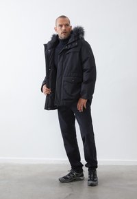 Parka negra aislada con capucha forrada de piel, cierre de cremallera, bolsillos frontales y un corte relajado. Llevada sobre un suéter oscuro y pantalones.