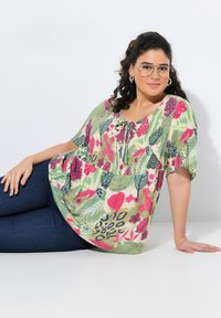Bluse mit Blumenmuster in Hellgrün, mit rosa und lila Akzenten, Rundhalsausschnitt mit Bindeband und kurzen Ärmeln. Kombiniert mit blauen Jeans.