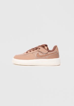 Brun børnesneaker med hvid sål, velcrolukning og syet Nike swoosh-logo på en ensfarvet grå baggrund.