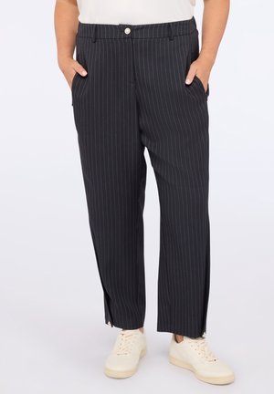 Czarne spodnie w prążki o prostych nogawkach z przednimi kieszeniami, noszone z białymi tenisówkami i białym topem, ręce w kieszeniach.