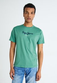 Zöld pamut póló rövid ujjakkal, elöl nyomott sötétkék "Pepe Jeans London" logóval. Világoskék farmernadrággal viselve.