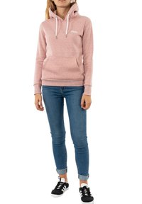 Sudadera con capucha en rosa claro con bolsillo tipo canguro, cordones y puños acanalados. Combinada con vaqueros azules y zapatillas negras. Ajuste y textura casual.