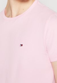 Helles rosa T-Shirt mit kurzen Ärmeln und kleinem gesticktem rechteckigen Logo in rot, weiß und schwarz auf der Brust, getragen von einer Person.