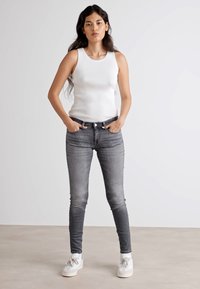 Biely rebrovaný tielko, šedé skinny džínsy a biele tenisky. Model stojí s rukami v vreckách na jednoduchej pozadí.
