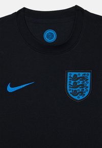 Camiseta negra de Nike con logo Swoosh azul, una etiqueta circular azul y un escudo de Inglaterra bordado en azul con tres leones y detalles florales.