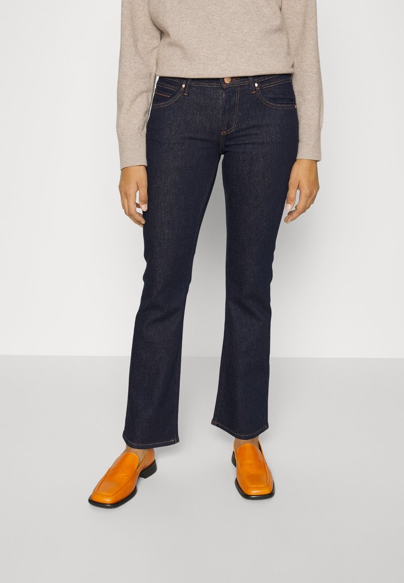 Jean flare bleu foncé avec une texture en denim lisse. Associés à des chaussures slip-on orange, avec un bout rond et une semelle noire.