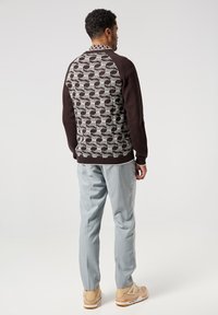 Hnědý pletený svetr s texturovaným šedým geometrickým vzorem na zadní straně, doplněný o světle šedé kalhoty a tanové tenisky.