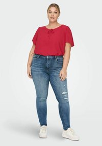 Červená blůza s krátkým rukávem a mašlí na výstřihu, spárovaná s opranými modrými skinny džíny a bílými teniskami. Viditelné texturované materiály.