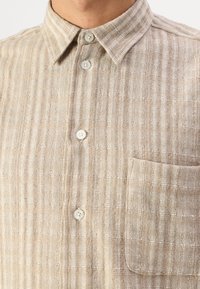 Camisa de lino en beige con un patrón texturizado y rayas; presenta un cierre de botones en la parte delantera y un bolsillo en el pecho con un solo botón.