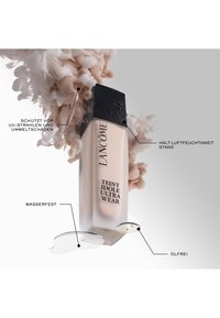 Lancôme fundamentflaske med frostet glasskropp, svart hette og tekstetiketter; omkranset av en virvlende, rosa væskebakgrunn.
