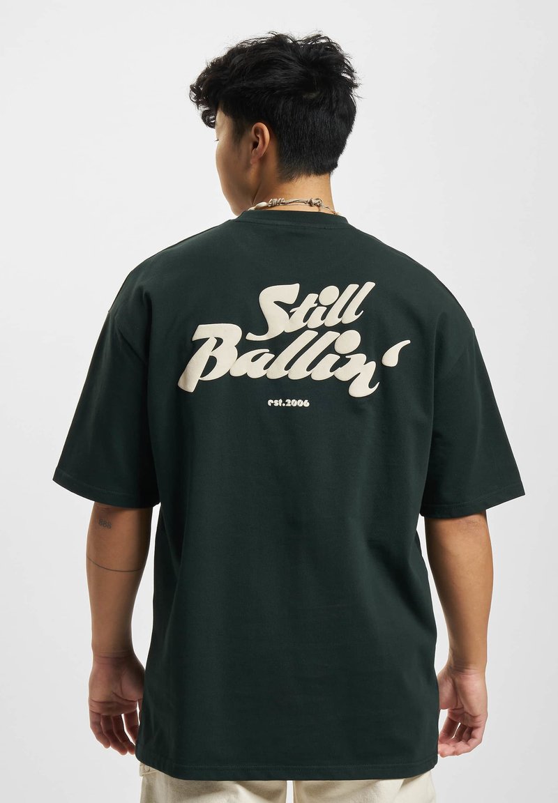 Tmavě zelené oversized tričko s krátkým rukávem, na kterém se nachází bílé nápisy "Still Ballin'" a detail "est. 2006" na zadní straně. Materiál: bavlna.