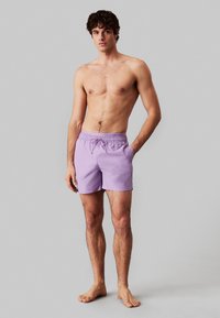 Costumi da bagno viola con vita elasticizzata e cordino. Realizzati in tessuto leggero, lunghi fino al ginocchio, caratterizzati da una superficie liscia.