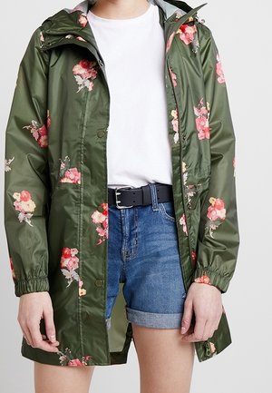Personne portant un imperméable vert à motifs floraux par-dessus un t-shirt blanc et un short en jean bleu retroussé avec une ceinture noire, debout devant un fond uni.