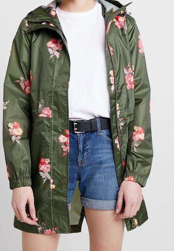 Personne portant un imperméable vert à motifs floraux par-dessus un t-shirt blanc et un short en jean bleu retroussé avec une ceinture noire, debout devant un fond uni.