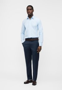 Moški stoji, oblečen v svetlomodro srajco, temnomodre hlače, rjav pas in rjavi škornji, na belem ozadju.