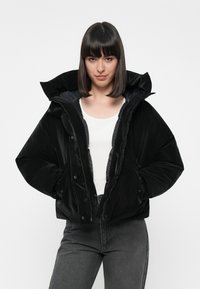 Giacca nera imbottita con colletto alto e cappuccio, realizzata in tessuto lucido. Presenta una chiusura con zip e tasche frontali, indossata sopra una camicia bianca.