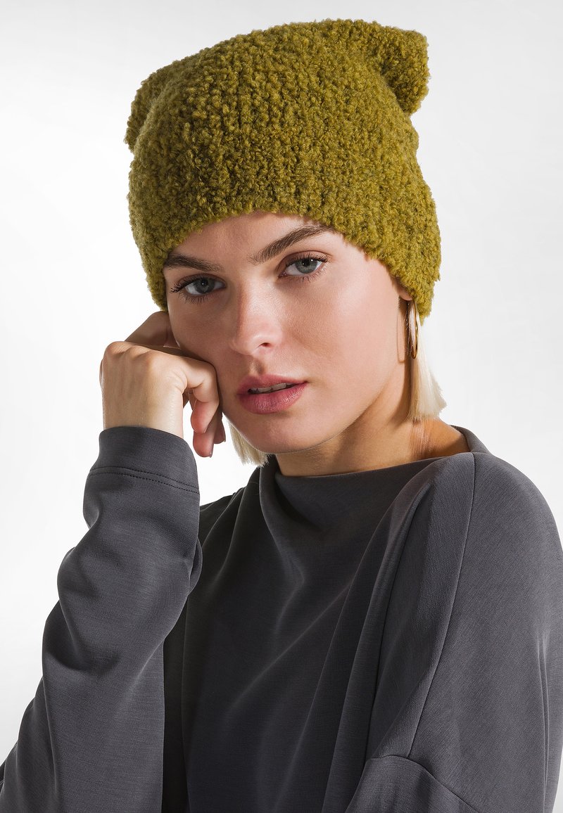 Mujer joven con el cabello rubio corto que lleva pendientes de aro dorados, un sombrero de punto verde oliva texturizado y una camiseta de manga larga gris oscuro, descansando su rostro sobre la mano.