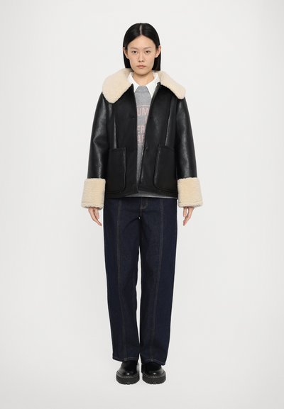 Giubbotto di pelle nera con collo e polsini in shearling sintetico color crema. Presenta due tasche frontali e un design casual, abbinato a pantaloni di denim.