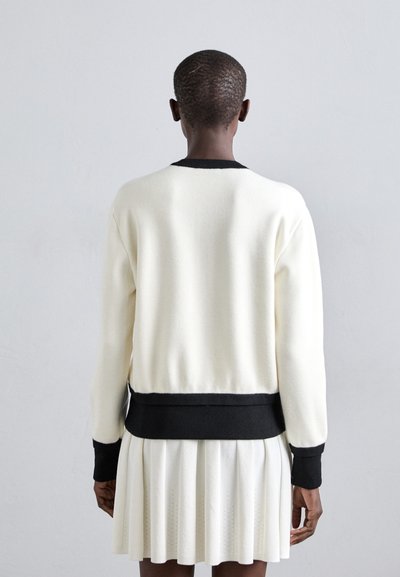 Pull blanc avec bord noir, texture douce, col rond et taille côtelée, associé à une jupe plissée blanche en tissu similaire.