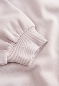 Gaiši rozā auduma paraugs ar ribotu aproci un savākta piedurknes detaļu, izceļot teksturētu adījuma virsmu. Gluda un maiga izskata.