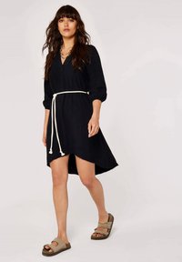 Schwarzes Kleid mit langen Puffärmeln, V-Ausschnitt und einem weißen Gürtel. Verfügt über einen asymmetrischen Saum und einen lässigen Schnitt. Getragen mit braunen Sandalen.