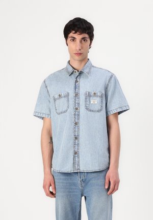 Hellblaues Denim-Hemd mit kurzen Ärmeln, Knopfleiste, zwei Brusttaschen und kontrastierenden Nähdetails. Glatte Textur.