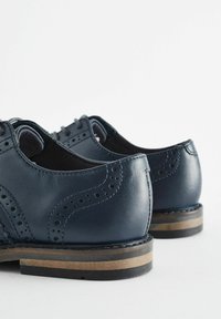 Marineblå læderdressko med brogue-detaljer, glat tekstur, rund tå og kontrasterende træ- og gummisål med en let hæl.