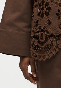 Χείρα με σκουρόχρωμο δέρμα κοντά σε καφέ ύφασμα με μεγάλο μανίκι και περίπλοκη λουλουδένια δαντέλα στη άκρη του ενδύματος.