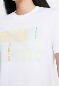 Model wearing a white t-shirt featuring a colorful Calvin Klein graphic print, focusing on brand names in various colors and styles.