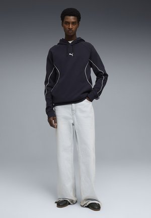 Joven de pie vistiendo una sudadera con capucha negra de Puma con ribetes blancos, jeans sueltos de lavado claro y zapatos negros, sobre un fondo gris liso.