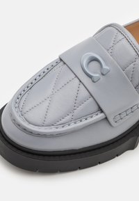 Mocassin en cuir gris avec texture matelassée et semelle en caoutchouc. Comprend une large bride et un accent de logo sur la partie supérieure.